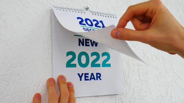 2021 'in Aralık sayfasındaki duvar takvimine göz atan erkek ellerinin yakın çekimi. Ardından yeni bir 2022 takviminin başlık sayfası.