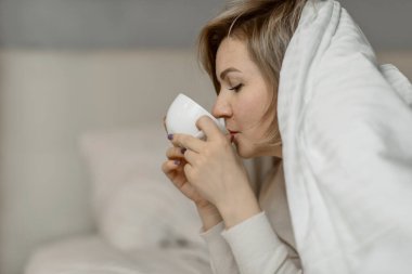 Orta yaşlı bir kadın, başının üzerinde battaniyeyle, sıcak bir bardak içecekle yatakta yüzüstü yatıyor.