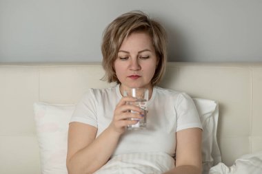 Orta yaşlı beyaz bir kadın, nemlendirilmiş güzel bir cilde sahip, elinde temiz bir bardak su tutuyor, yatağında oturuyor. Sağlık ve güzellik hizmetleri kavramı
