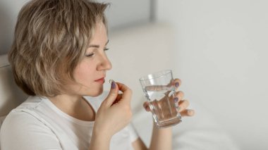 Orta yaşlı beyaz bir kadın, nemlendirilmiş güzel bir cilde sahip, elinde bir bardak temiz su ve vitaminle yatakta oturuyor. Sağlık ve güzellik hizmetleri kavramı