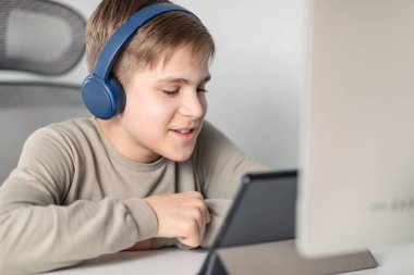 Mavi kulaklıklı sarışın genç bir çocuk evde monitörün önündeki masada oturuyor ve online bir toplantıya katılıyor.