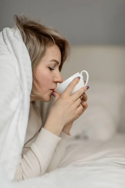 A middle-aged woman is lying on her stomach in bed, covered with a blanket over her head, with a cup of warm drink