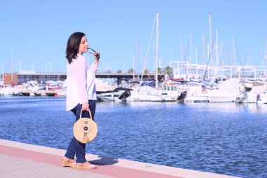 Tüm vücudu kapalı, beyaz tenli orta yaşlı kadın rıhtımda güneş gözlüklü, tekne arka planında poz veriyor. Akdeniz 'de yaz mevsiminde yaşam tarzını kavrayın