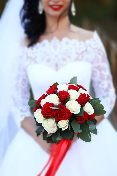 Wedding Bouquet in the Hands of the Bride. The Bride Holds a Bouquet of Flowers from Roses Outdoors. Stylish Wedding Bouquet on the Background of the White Dress of the Beautiful Bride.