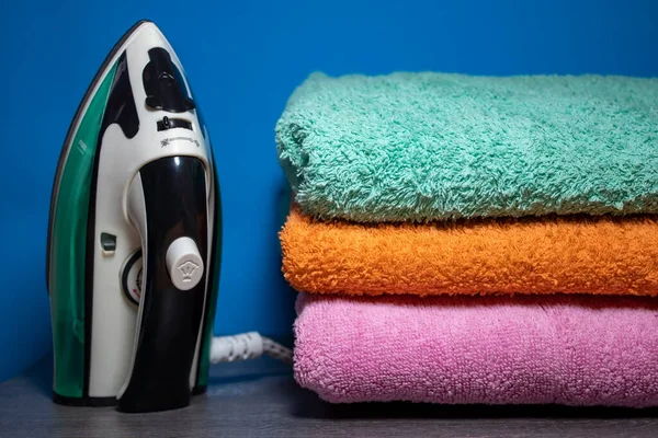 New Iron and Ironing Towels on the Table. Iron Irons linen. Pile of Clean Stacked Home Textile Items Near Blue Wall. Cozy House, Laundry and Ironing. Iron and Ironed Linen on a Wooden Surface.