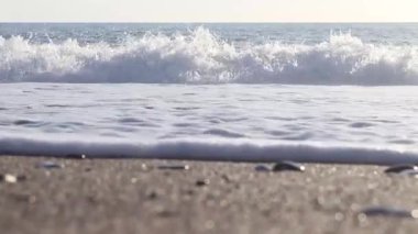 Siyah, beyaz ve bej renkli deniz taşlarının yakın çekim dokusu. Kayalık bir sahilde deniz dalgaları köpüğü. Taşların arka planı pürüzsüz hareket ediyor. Çok renkli deniz taşları güneşte parıldıyor. 