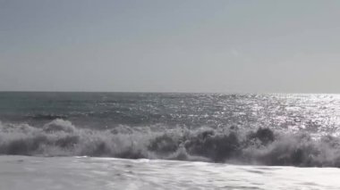  Kayalık bir sahilde deniz dalgaları köpüğü. Taşların arka planı pürüzsüz hareket ediyor. Çok renkli deniz taşları güneşte parıldıyor.