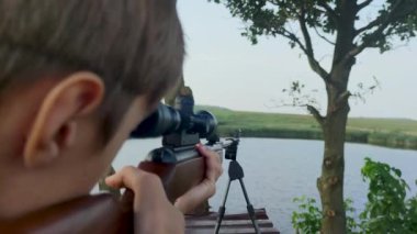The Guy Makes a Shot with an Air Rifle. The Sight is Aimed at the Mark Behind the Lake. The Guy Shoots Outside the City on a Warm Summer Day.