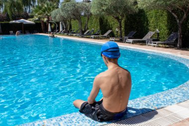 A Young Boy is Sitting Near the Pool. Empty Space for Text. A Boy Sits by the Pool and Prepares to Dive and Swim in the Clear Blue Water in the Open Air Street of Calabria, Italy.