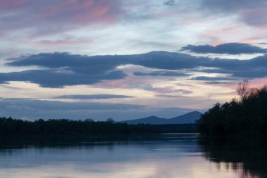 Alacakaranlıkta nehir ve dağ siluetinin manzarası, ormanlık kıyısı olan Sava nehri ve mavi saat boyunca gökyüzünde bulutlar ve bulutlarda mor parlayan Motajica dağı.