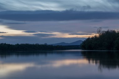 Alacakaranlıkta nehir ve dağ siluetinin manzarası, ormanlık kıyısı olan Sava nehri ve mavi saat boyunca gökyüzünde bulutların olduğu Motajica dağı manzarası