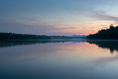 Su yüzeyinde sisli nehir manzarası ve alacakaranlıkta uzak dağ, simetrik yansıma ve pastel renkler