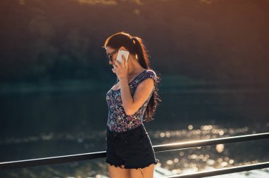 Çekici hippi kız açık havada iskelede 4G bağlantısı olan yeni cep telefonuyla postaları kontrol ediyor, pozitif kadının nehir kenarında dinlenirken modern cep telefonundan mesaj gönderdiğini görüyor..