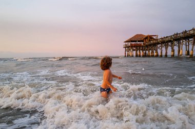 Gün batımında dalgalara atlayan bir çocuk. Mutlu çocuk yaz plajında hayatın tadını çıkarıyor. Güneşli bir günde denizdeki dalgaların arasında küçük bir çocuk.