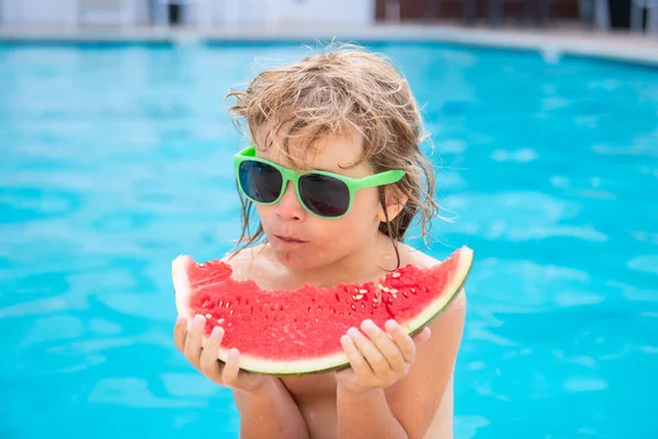 Child eating watermelon near swimming pool during summer holidays. Kids eat fruit outdoors. Healthy food for children. Little boy playing in pool with a slice of water melon.