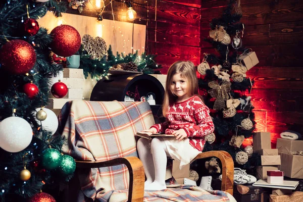 Niña de Navidad con un libro en sus manos sentada junto al árbol de Navidad. Feliz niño lindo en el sombrero de Santa con regalo tienen una Navidad. Año nuevo concepto de Navidad. —  Fotos de Stock