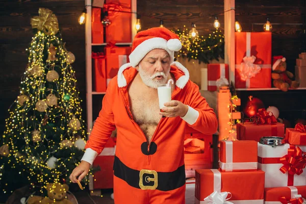 サンタは家でクッキーと牛乳を選んでいます。クリスマスカードを。ミルクとジンジャーブレッドクッキークリスマスライトの背景に対するサンタ. — ストック写真