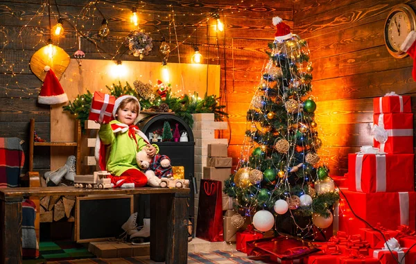 Mutlu küçük çocuk Noel ağacının yanında oyuncaklarla oynuyor ve Noel hediyesini kameraya gösteriyor. Küçük çocuk Noel Baba kıyafeti giyiyor. Tatil kavramı. — Stok fotoğraf