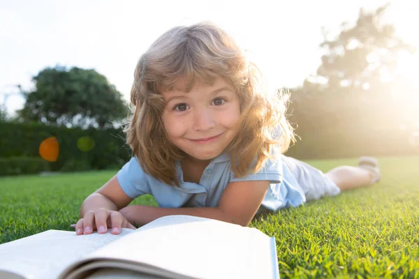 Roztomilý chlapec čte knihu ležící na trávě. Dítě čte knihu v letním parku. — Stock fotografie