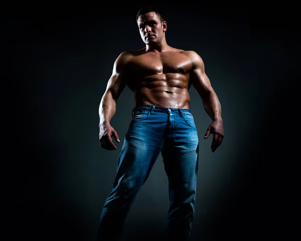 Sexy beau bronzé homme caucasien en jeans, debout torse nu, montrant six pack abdos abdominaux, ont un corps parfait, fond noir foncé. — Photo