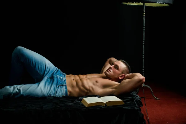 Een gespierde en atletische ontklede man met een kale romp die een boek leest. Sexy kerel in de slaapkamer. — Stockfoto