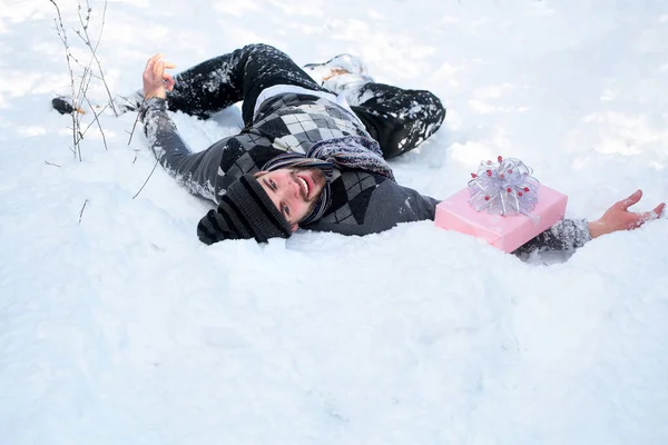Homme fou avec boîte cadeau couché sur la neige. Salutations de saison et cadeaux de Noël. Macho gay souriant dans chapeau, pull et écharpe le jour de l'hiver — Photo