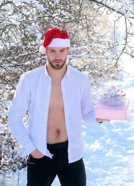 Sexy man in kerstmuts met kistje in sneeuwhout. Macho met naakte romp in open shirt op winterdag. Seizoen groeten en kerstcadeaus. Vakantie viering concept. Vrolijk kerstfeest en gelukkig nieuw — Stockfoto