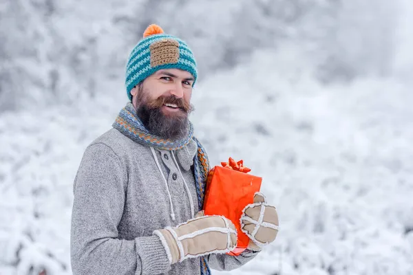 Zimní muž. Vánoční šťastný muž s vousy držet dárkovou krabici. Hipster santa v chladném zasněženém zimním lese — Stock fotografie