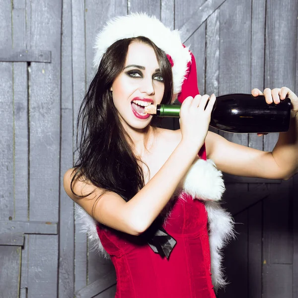 Woman pull out cork with teeth. Attractive woman in Santa Claus corset and red hat in studio on wooden background with corked wine bottle — Stock Photo, Image