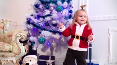 Kırmızı Noel Baba süveteri ve Noel Baba şapkasıyla Noel ağacında gülümseyen küçük çocuk..