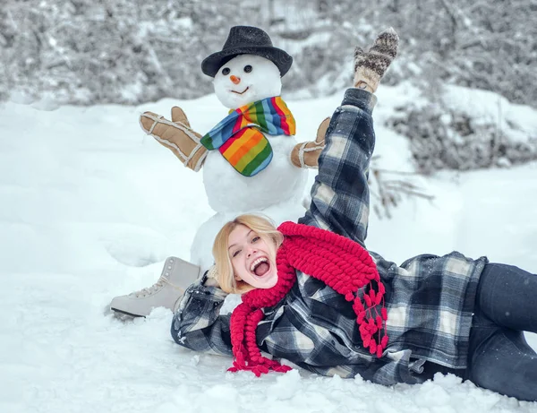 Zimní žena. Zdravím sněhuláka. Sněhulák a legrační modelka stojící v zimním klobouku a šále s červeným nosem. Roztomilý sněhulák na zasněžené vesnici. — Stock fotografie
