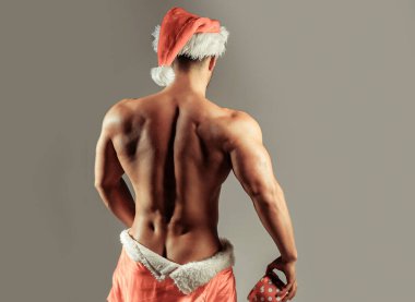 Seksi noel baba. Şapkalı kaslı, seksi vücutlu, arkasında duran, kırmızı Noel Baba ceketinin altında kalçalarını saklayan, gri arka planda hediyesini tutan adam.