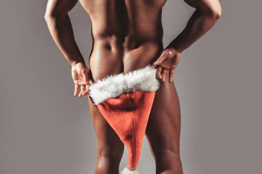 Sexy muscular gay man hiding buttocks butt, ass under red santa claus hat. Winter holidays celebration on grey