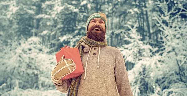 Zimní muž s vousy drží dárkovou krabici. Hipster Santa v chladném zasněženém zimním lese. Vousatý muž v Santa klobouku na Nový rok — Stock fotografie