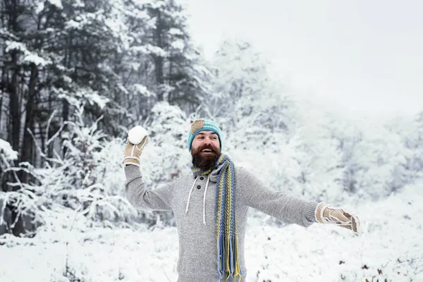 Hipster em casaco, chapéu, cachecol, barba quente no inverno. Homem barbudo sorrir com bola de neve na floresta nevada. Papai Noel barbudo — Fotografia de Stock