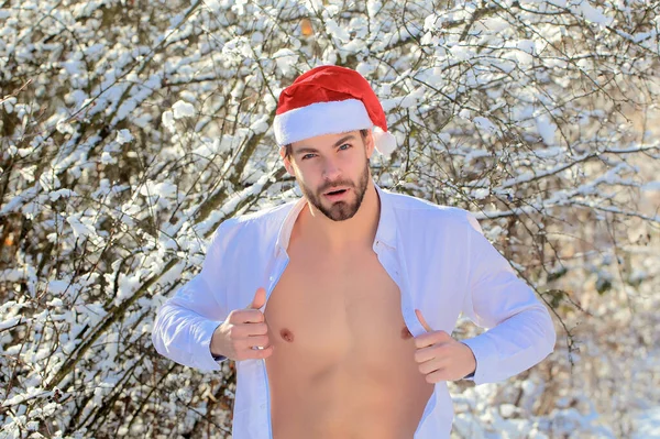 Homo met gespierde romp in de winter buiten. Man uitkleden shirt in de winter. Macho in kerstmuts in sneeuwhout — Stockfoto