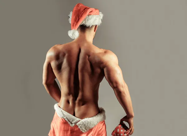 セクシーなサンタさん。Muscular man in hat with sexy body standing with back, hides buttocks under red santa claus coat, holds gift on grey background — ストック写真