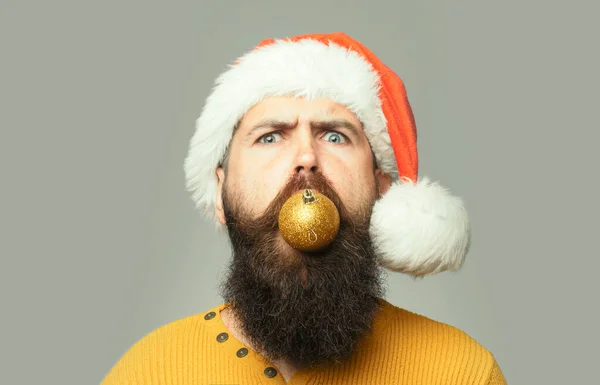 Galning man med långt skägg och mustasch på allvar ansikte med julkula i munnen på grå bakgrund. Närbild rolig ansikte porträtt — Stockfoto