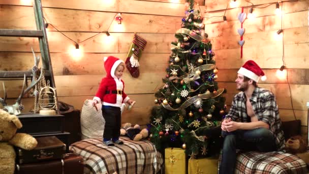 Bébé enfant drôle avec jeune père ou frère à la maison sur fond d'arbre de Noël. Petit enfant au chapeau de Père Noël, Nouvel An. Soirée d'hiver chaleureuse à la maison. — Video