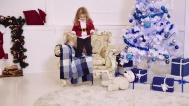 Noel ağacı arka planında evdeki kanepeden atlayan harika bir çocuk. Ağır çekim. Noel Baba şapkalı küçük çocuk komik bir atlayış.