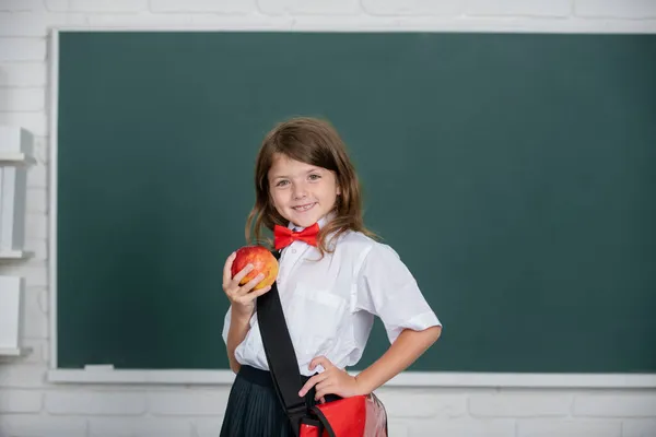Glad leende preteen flicka, unge skolflicka med bär schhol uniform hålla ryggsäck på svarta tavlan bakgrund tittar på kameran. Begreppet utbildning, lärande och barn. — Stockfoto