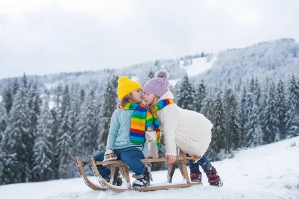 Діти цілуються мило. Щасливий маленький хлопчик і дівчинка катаються взимку і цілуються. Діти катаються на санях. На відкритому повітрі діти розважаються на Різдвяні канікули . — стокове фото