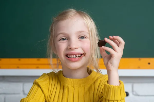 クラスルームのテーブルのデスクトップに座って、チョコレートを食べて笑顔の小さな幸せな女の子のクローズアップ肖像画。小さな面白い学校の女の子顔. — ストック写真