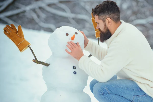 Bonhomme de neige et drôle homme barbu dans la neige. Joyeux beau jeune hipster s'amuser avec bonhomme de neige en hiver. — Photo