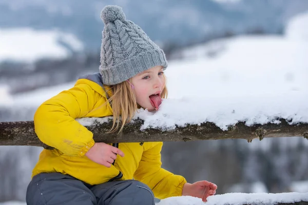 冬の女の子は屋外で雪を食べる。面白いクリスマスの子供の休日。風邪やインフルエンザの概念。舌で雪を舐めなさい。雪の日に暖かい冬服を着た子供たち. — ストック写真