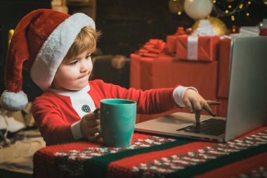 Çocuklar için online Noel alışverişi. Küçük çocuk dizüstü bilgisayarının yanında oturan Noel Baba kıyafetleri giyiyor. Not defteri kullanarak Noel Baba yardımcısı.