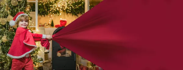 Niño Santa con bolsa de regalos, pancarta aislada sobre fondo rojo con espacio para copiar. Ayudante de Santa celebración con bolsa roja con regalos en el árbol de Navidad. Ayudante de Santa Claus llevando una gran bolsa llena de regalos — Foto de Stock