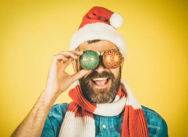 Julhipster glad leende i Santa hatt och halsduk. Man täcka ögon med xmas bollar på gul bakgrund. Semesterdekorationer — Stockfoto