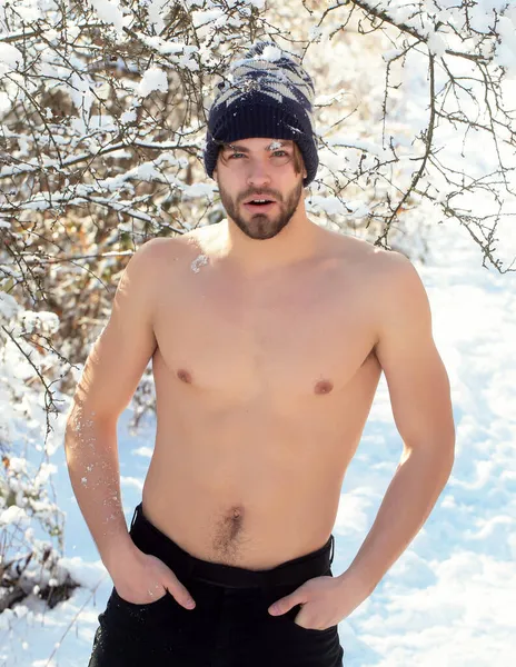 Ernstige knappe jonge man of sexy gespierd gay met kale romp en naakte borst lichaam in de winter hoed buiten in bomen met sneeuw — Stockfoto