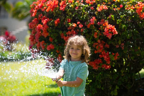 Glad pojke häller vatten från en slang. Barnvattning blommor i trädgården. Trädgårdsskötsel. — Stockfoto